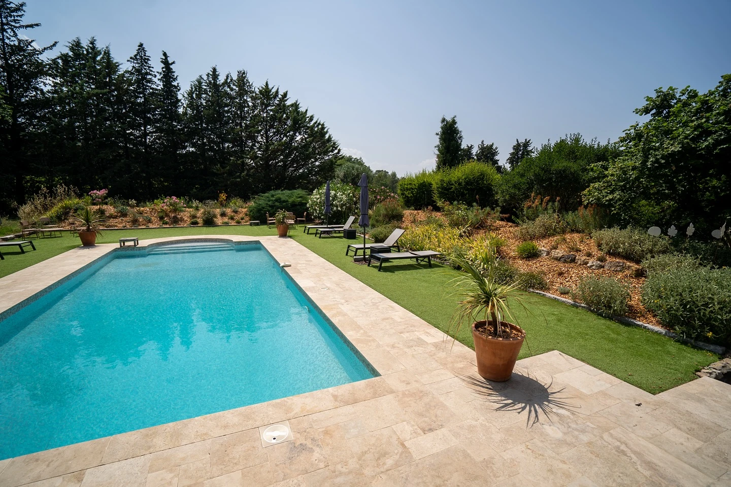 Piscine extérieure avec escalier immergé en pierre naturelle, entourée de végétation méditerranéenne incluant palmiers et lauriers-roses en fleurs