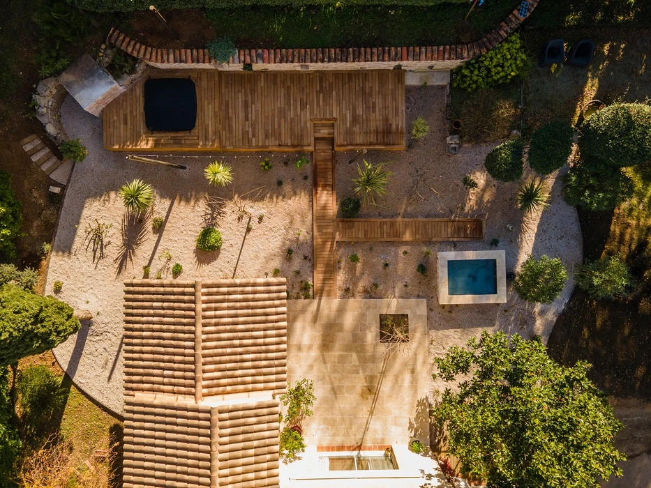 Jardin méditerranéen ensoleillé avec olivier centenaire au premier plan, pelouse synthétique verte, muret en pierre calcaire taillée