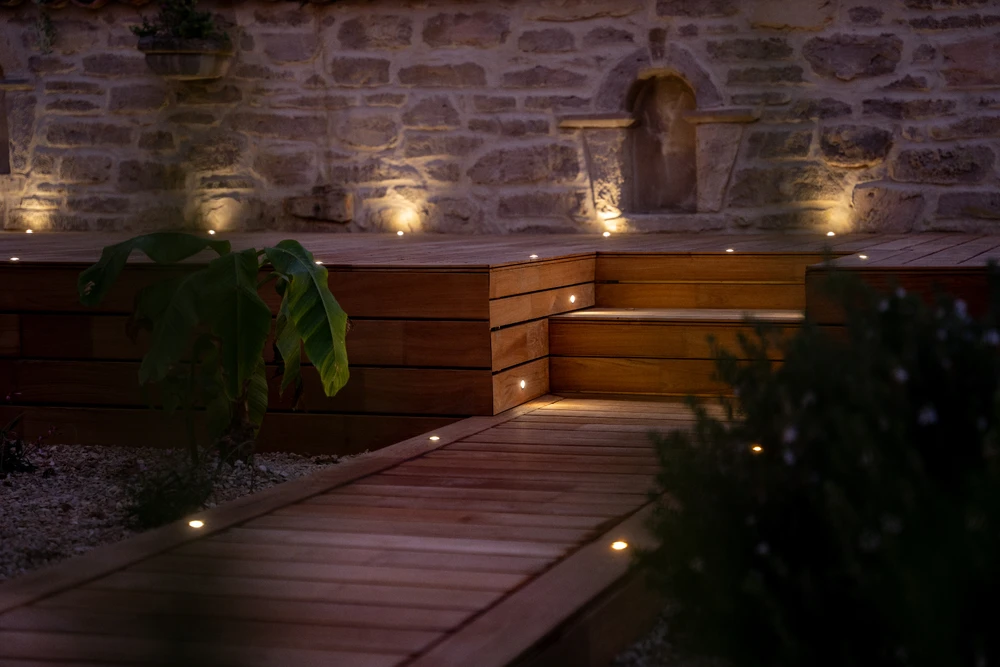 Jardin résidentiel ensoleillé, pelouse verte tondue, petit bâtiment annexe blanc avec toit en tuiles canal terre cuite, clôture à mailles losangées galvanisée sur poteaux verts, arbuste sec, pin parasol, chaise de jardin plastique noire, fond de végétation dense